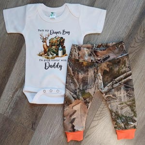 Może przedstawiać: Kremowy zestaw dla niemowląt składający się z body i spodni w kamuflażu. Body posiada grafikę jelenia z plecakiem i napisem "Pack my Diaper Bag, I'm going huntin' with Daddy." Spodnie mają pomarańczowe mankiety.