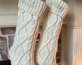May include: Two cream-colored cable knit Christmas stockings hang from a white ribbon. The top cuff of each stocking is embroidered with a name in silver thread: "Maddie" and "Courtney". The stockings are displayed in front of a brick fireplace.