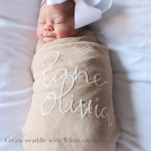 K&ouml;nnte beinhalten: Eine cremefarbene Wickeldecke mit wei&szlig;er Stickerei, auf der "Baby Olivia" steht.