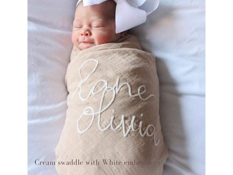 K&ouml;nnte beinhalten: Eine cremefarbene Wickeldecke mit wei&szlig;er Stickerei, auf der "Baby Olivia" steht.