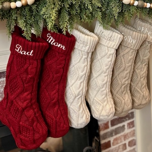 May include: Six cable-knit Christmas stockings in varying colors hang from a garland. Two are red with "Dad" and "Mom" embroidered on them. The others are cream and tan, each with a name embroidered in a matching color. The stockings are displayed near a brick fireplace.