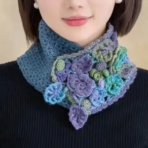 May include: A blue and green crocheted scarf with flower embellishments. The scarf is worn around the neck of a person wearing a black sweater.