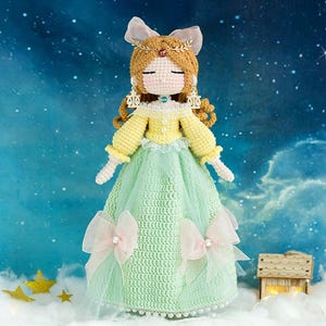 May include: A handmade crochet doll with a yellow top, a light green skirt, and a white bow. The doll has brown hair, a gold crown, and is set against a starry background. The doll is approximately 25 cm tall.