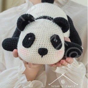 young girl sporting cute lying panda amigurumi/toy/doll, panda on both hands of girl facing camera, panda in lying position with his butt rising up high, panda is crocheted with plushie yarn, panda having round face and nose, with indoor background