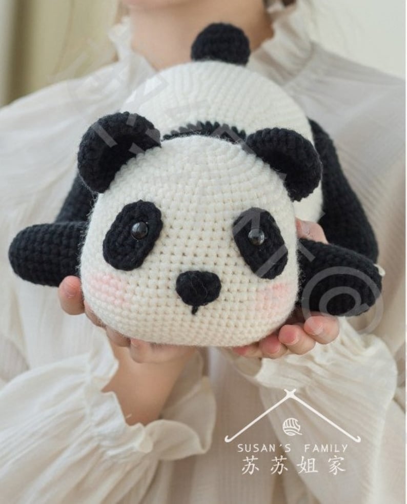 young girl sporting cute lying panda amigurumi/toy/doll, panda on both hands of girl facing camera, panda in lying position with his butt rising up high, panda is crocheted with plushie yarn, panda having round face and nose, with indoor background