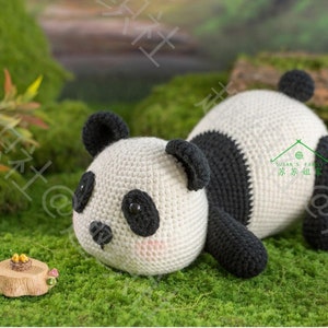 45 degree left front view of panda amigurumi/plushie toy/doll lying with chin on grassy ground, panda watching sth. tiny in front him, his limb, tummy and chin all resting on ground, with hip rising up high in air, with garden background