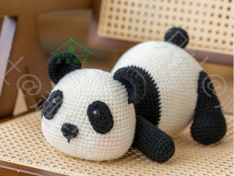 left side front view of crochet panda amigurumi/plushie lying on tummy on ratten weaven chair, panda bear in black and white, round face, round butt and tail sticking up in air , short arms and legs, with indoor background