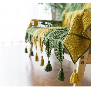 45 diagonal front view of couch cover, cover in green and yellow colors in Victorian-style granny square tile pattern, cover edge is triangle patches with tassels, with indoor background
