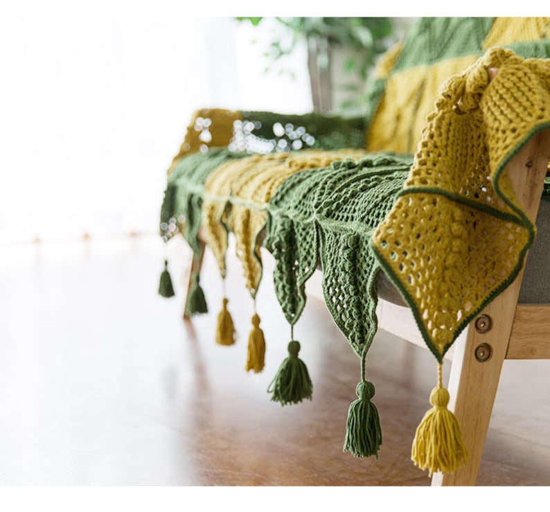 45 diagonal front view of couch cover, cover in green and yellow colors in Victorian-style granny square tile pattern, cover edge is triangle patches with tassels, with indoor background