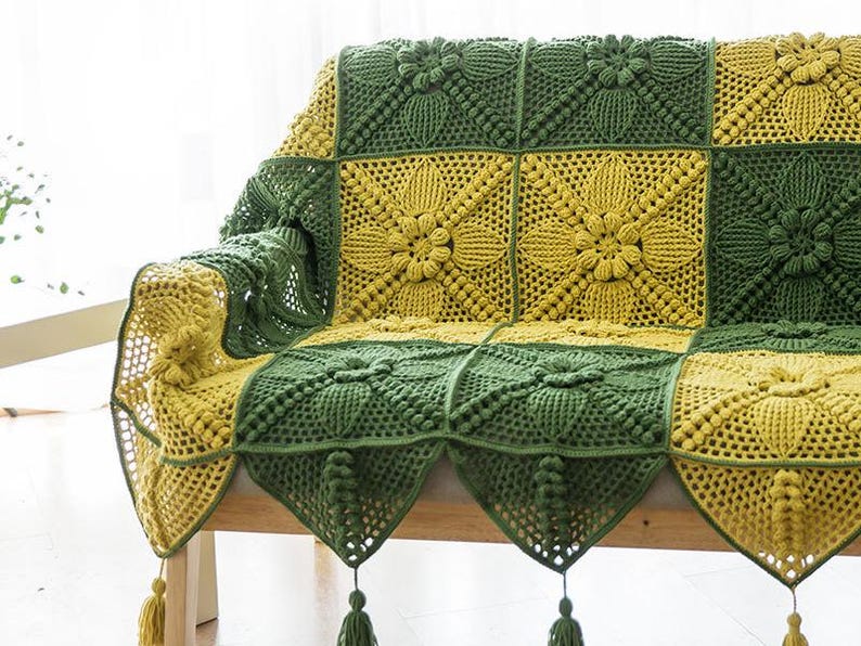view of left sise of couch or sofa covered with crochet sofa cover of couch cover, cover is in green and yellow or ginger color, with granny square pattern with a lot of poocorn crochet, edge of blanket is in triangle shape, decorated with tassel.