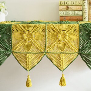close up details of granny square table cloth, cloth is made of yellow (ginger) and dark green granny square and triangle blocks, and decorated with tassels, granny square has floral pattern