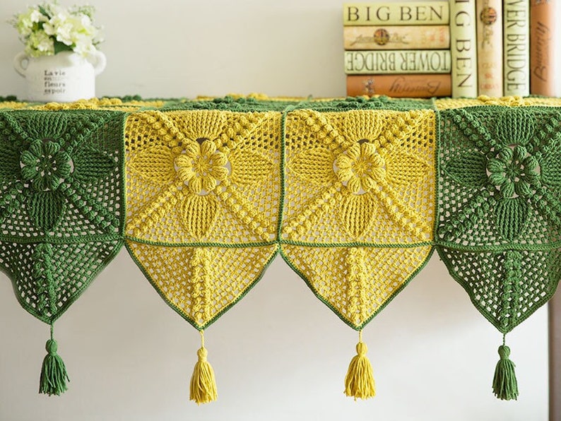 close up details of granny square table cloth, cloth is made of yellow (ginger) and dark green granny square and triangle blocks, and decorated with tassels, granny square has floral pattern