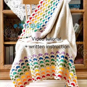 May include: A crocheted rainbow blanket draped over a wooden chair. The blanket features a colourful, scalloped border and a white centre. The text "Video tutorial + written instruction" is visible on the blanket.