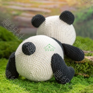 back view of crochet panda lying on green grass landscope on tummy, panda having white round fat, body and butt, short round black tail, black short fat limbs and ears, panda looking up with tummy and limbs lying on ground, with garden background