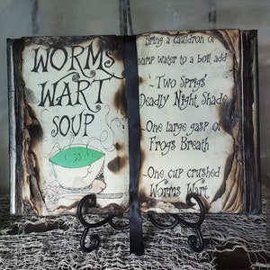 May include: A vintage-style book with a burnt-edge design, titled "Worms Wart Soup." The book is open to a page with a recipe for the soup, including ingredients like "Deadly Night Shade" and "Frogs Breath."