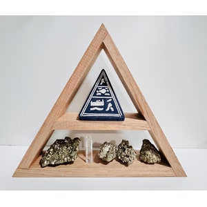 Könnte beinhalten: Ein dreieckiges Holzregal mit zwei Ablagen. Die obere Ablage hat eine blaue Pyramide mit weißen Markierungen. Die untere Ablage hat vier goldfarbene Steine und einen klaren Kristall.