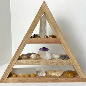 Puede incluir: Una estantería triangular de madera con tres niveles, que muestra una variedad de cristales y piedras, incluyendo cuarzo transparente, amatista, ojo de tigre y citrino.