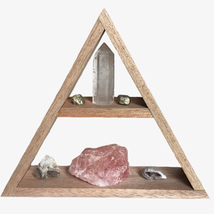May include: A wooden triangular shelf with two shelves, displaying various crystals and stones. The top shelf has a clear quartz crystal and two gold pyrite stones. The bottom shelf has a large rose quartz crystal, a small clear quartz crystal, and a gray agate stone.