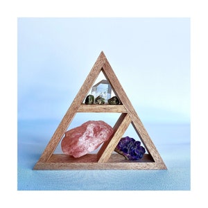 May include: Wooden triangular crystal shelf with four compartments. The top compartment has a clear crystal, the middle compartment has a pink crystal, the bottom left compartment has a green crystal, and the bottom right compartment has a purple crystal.