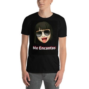 May include: Black t-shirt featuring a graphic of an emoji with sunglasses and a tongue sticking out. The text "Me Encantas" is printed in white and red below the emoji. The shirt is worn by a person.