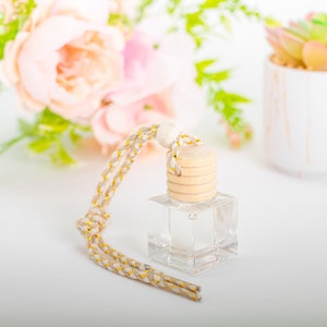 Op de afbeelding: Een heldere glazen fles met een houten deksel en een beige en gouden touwtje. De fles is gevuld met een heldere vloeistof en hangt aan het touwtje.