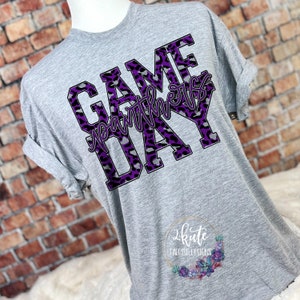 May include: Gray t-shirt with a purple and black leopard print design that says "GAME DAY" with the word "GAME" in all caps and the word "DAY" in all caps. The word "DAY" is in a larger font than the word "GAME". The words "with the" are written in a smaller font and are in between the words "GAME" and "DAY".