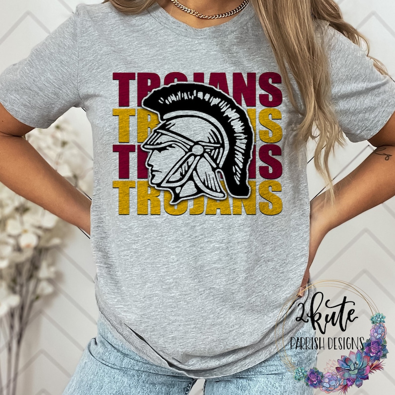 K&ouml;nnte beinhalten: Ein graues T-Shirt mit einem Grafikdesign eines schwarz-wei&szlig;en r&ouml;mischen Helms mit dem Wort "TROJANS" in Rotbraun und Gold wiederholt.