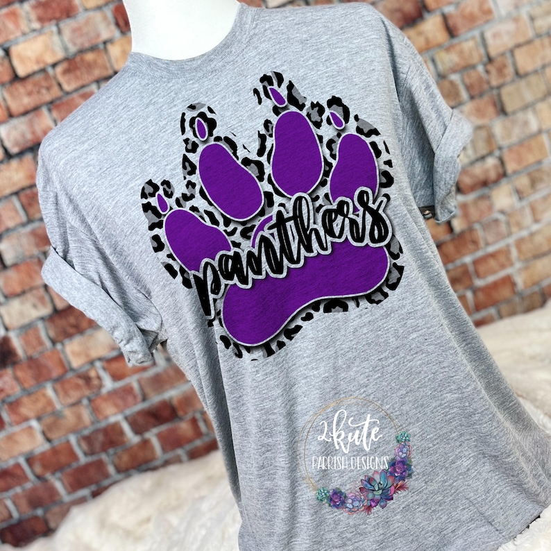 May include: Gray t-shirt with a purple paw print design and the word "Panthers" in black lettering. The paw print is on a leopard print background.
