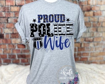 May include: Gray t-shirt with the text "Proud Police Wife" in black and blue lettering with a blue and white American flag design and a blue police badge.