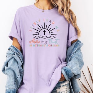 May include: A lavender t-shirt with a graphic of a sunrise with a cross and the text "Spirit Lead Me where my trust is without borders".