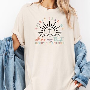 May include: A beige t-shirt with a graphic design of a sunrise with a cross and the text "Spirit Lead Me where my trust is without borders".