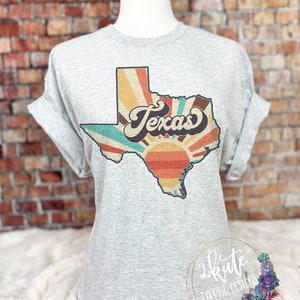 May include: A grey t-shirt featuring a vintage-style graphic of the state of Texas. The graphic has a sunburst design with the word "Texas" in a retro font.