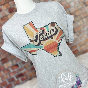 May include: A grey t-shirt featuring a vintage-style graphic of the state of Texas with a sunburst design and the word "Texas" in a retro font.