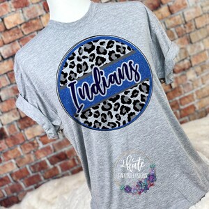 Pu&ograve; includere: Maglietta grigia con un design circolare blu con stampa leopardata grigia e nera e la parola "Indians" in lettere glitter blu.