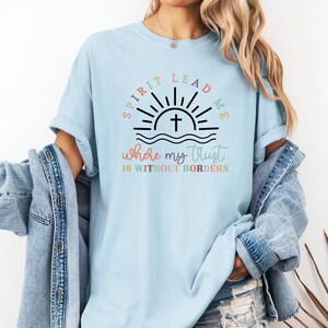 May include: A light blue t-shirt with a graphic design of a sunrise with a cross and the text "Spirit Lead Me Where My Trust Is Without Borders". The text is in rainbow colors.