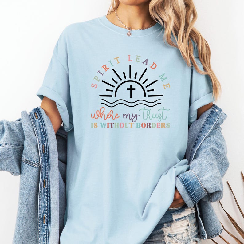May include: A light blue t-shirt with a graphic design of a sunrise with a cross and the text "Spirit Lead Me Where My Trust Is Without Borders". The text is in rainbow colors.