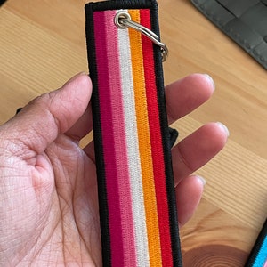 Op de afbeelding: Een zwarte sleutelhanger met een ontwerp van een lesbische pride-vlag. De vlag heeft roze, witte, oranje en rode strepen. De sleutelhanger heeft een zilveren ring.