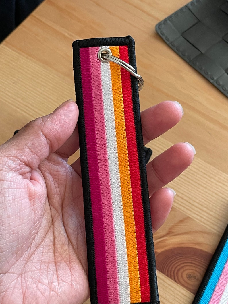 Op de afbeelding: Een zwarte sleutelhanger met een ontwerp van een lesbische pride-vlag. De vlag heeft roze, witte, oranje en rode strepen. De sleutelhanger heeft een zilveren ring.