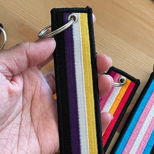 Op de afbeelding: Een zwarte, paarse, witte en gele gestreepte sleutelhanger met een zilveren ring. De sleutelhanger is gemaakt van stof en heeft een gestikt ontwerp.