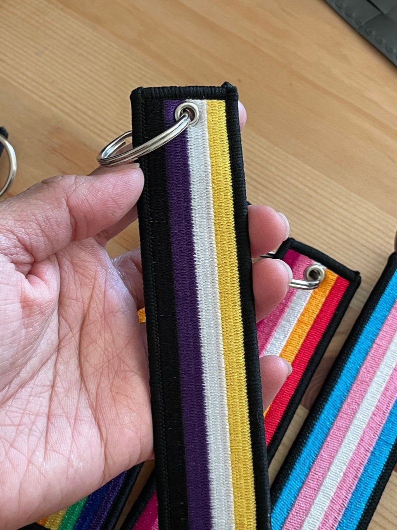 Op de afbeelding: Een zwarte, paarse, witte en gele gestreepte sleutelhanger met een zilveren ring. De sleutelhanger is gemaakt van stof en heeft een gestikt ontwerp.