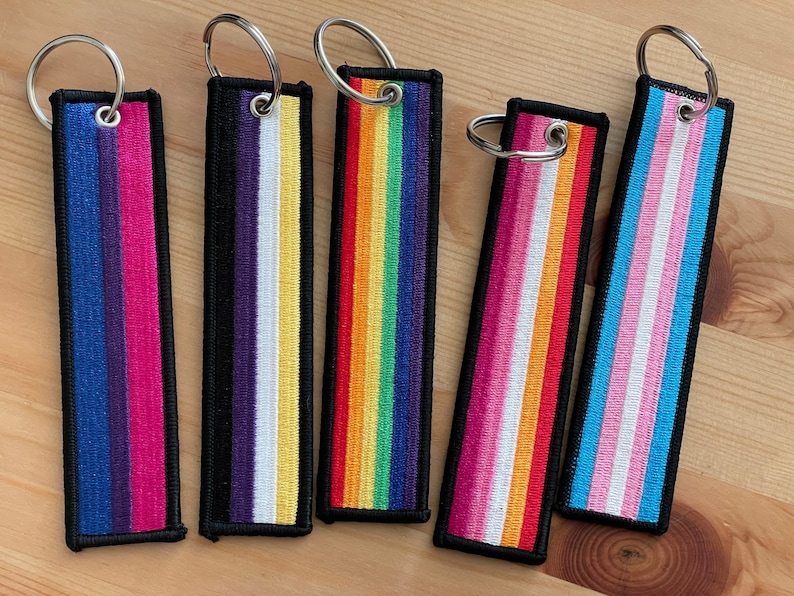 Op de afbeelding: Vijf verschillende sleutelhangers met pride-vlaggen, zwarte randen en zilveren sleutelringen. De vlaggen zijn biseksueel, non-binair, regenboog, transgender en lesbisch.