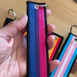 Op de afbeelding: Een set van vier sleutelhangers met verschillende regenboogvlagontwerpen. De sleutelhangers zijn gemaakt van zwart stof met gekleurde strepen. De vlaggen zijn biseksueel, transgender, lesbisch en regenboog.