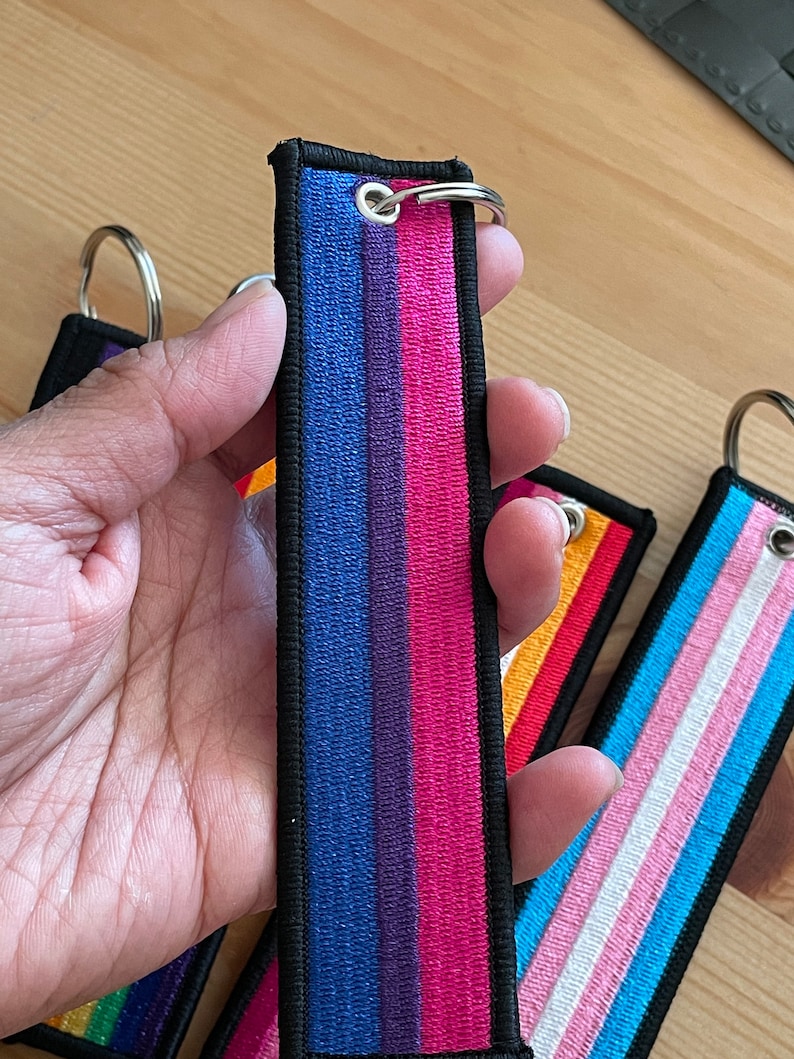 Op de afbeelding: Een set van vier sleutelhangers met verschillende regenboogvlagontwerpen. De sleutelhangers zijn gemaakt van zwart stof met gekleurde strepen. De vlaggen zijn biseksueel, transgender, lesbisch en regenboog.