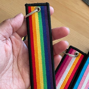 Op de afbeelding: Een set van drie regenboogkleurige sleutelhangers met zwarte randen en zilveren ringen. De sleutelhangers hebben verschillende ontwerpen van regenboogvlaggen, waaronder de traditionele regenboogvlag, de transgendervlag en de biseksuele vlag.