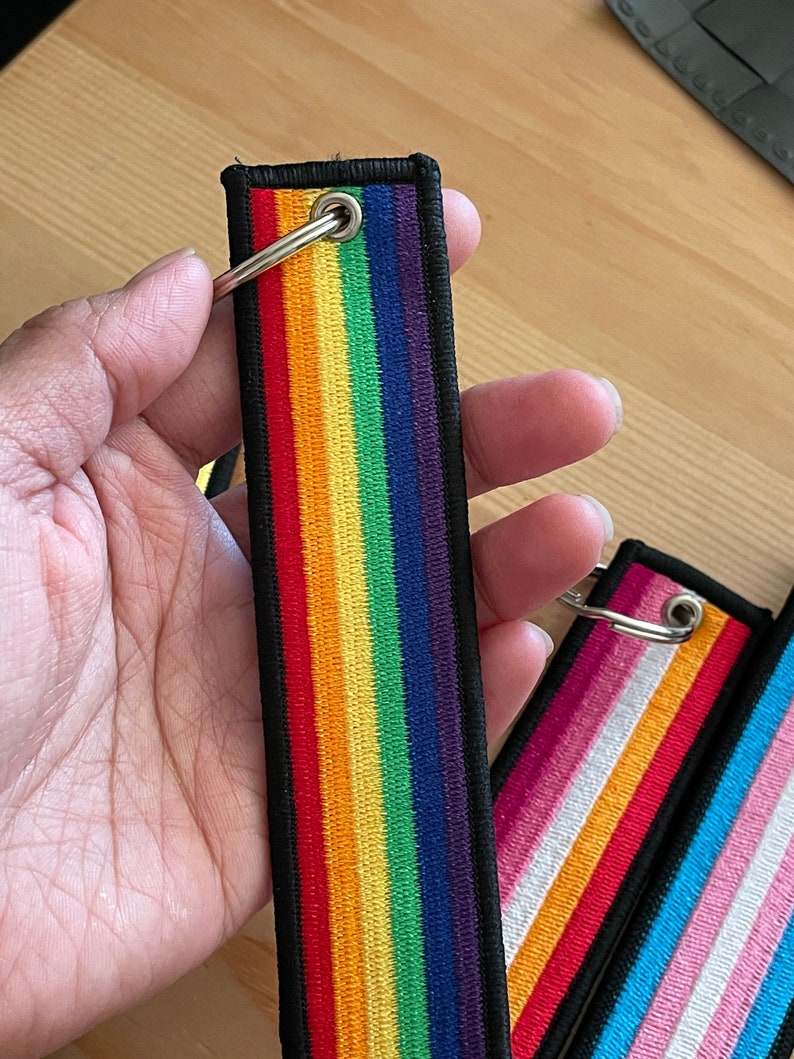 Op de afbeelding: Een set van drie regenboogkleurige sleutelhangers met zwarte randen en zilveren ringen. De sleutelhangers hebben verschillende ontwerpen van regenboogvlaggen, waaronder de traditionele regenboogvlag, de transgendervlag en de biseksuele vlag.