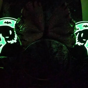K&ouml;nnte beinhalten: Ein Paar schwarze, im Dunkeln leuchtende Mickey-Mouse-Ohren mit gr&uuml;nem Leuchten. Die Ohren zeigen eine Silhouette eines Geistes mit Flederm&auml;usen.