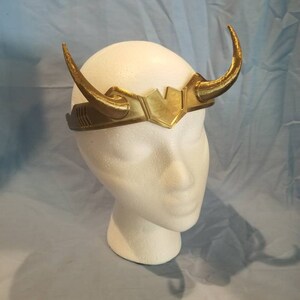 May include: A gold-colored headband with two horns, resembling a crown. The headband is made of plastic and has a textured surface.