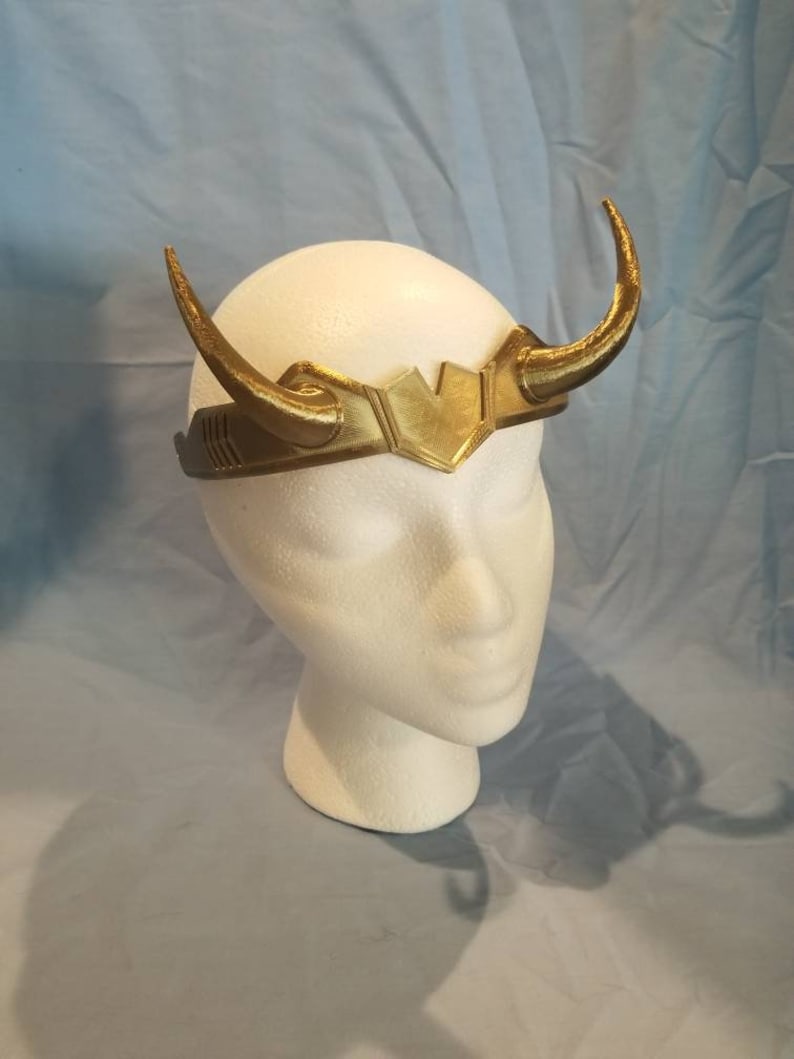 May include: A gold-colored headband with two horns, resembling a crown. The headband is made of plastic and has a textured surface.