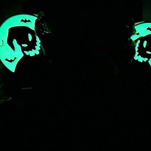 K&ouml;nnte beinhalten: Zwei im Dunkeln leuchtende gr&uuml;ne und schwarze Halloween-Ohrringe. Jeder Ohrring zeigt einen Halbmond mit der Silhouette einer gruseligen Figur mit einem zahnigen Grinsen und Fledermausfl&uuml;geln.