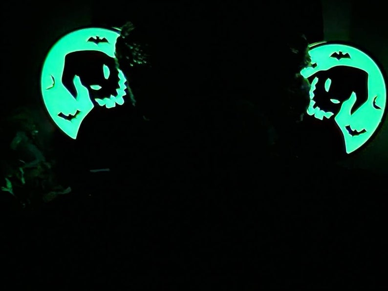 K&ouml;nnte beinhalten: Zwei im Dunkeln leuchtende gr&uuml;ne und schwarze Halloween-Ohrringe. Jeder Ohrring zeigt einen Halbmond mit der Silhouette einer gruseligen Figur mit einem zahnigen Grinsen und Fledermausfl&uuml;geln.